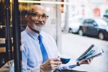 Beyaz gömlekli, kısa saçlı, kahve fincanı ve gazete kupasıyla caddenin yakınındaki sokağa bakan yaşlı bir adamdan.