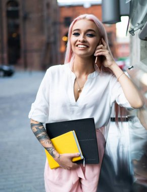 Sokakta cep telefonuyla konuşurken elinde not defterleri olan ve kameraya bakan aktif ve modaya uygun genç bir bayan.