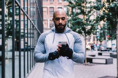 Ciddi, kel, sakallı, spor giyimli Amerikan erkek. Akıllı telefon kullanıyor ve modern binanın yanında sokak ortasında müzik dinliyor.