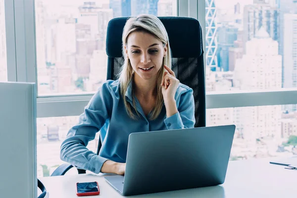 Confident young woman watching laptop working with gadgets at computer desk in contemporary office with cityscape behind window on background