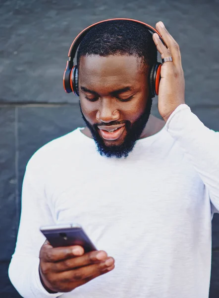 Dark skinned hipster guy updating music app on mobile phone to download audio via 4G internet connection, young man listening music playlist from cellular application via bluetooth headphones