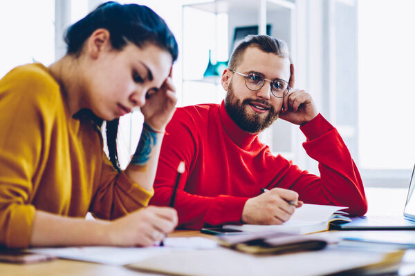 Young hipster guy in spectacles pondering on creative idea cooperating with female colleague noting information, clever student sitting at desktop learning and making researches for homework