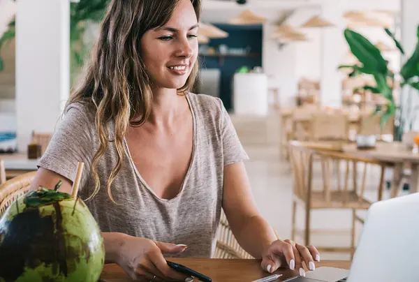 Young millennial woman checking setting on laptop device sitting at table with coconut cocktail and using cafeteria web connection for online communication, concept of digital nomad lifestyle