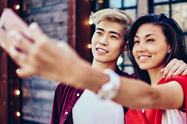 Happy chinese couple in love standing together outdoors on background with modern cafe and making photo on front camera of smartphone.Positive asian friends taking selfie picture on cellular