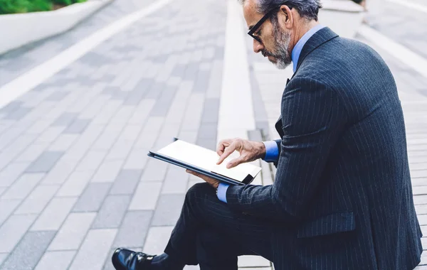 Side view of concentrated middle aged man in business suit and glasses with legs crossed working on tablet while sitting on bench against street of New York