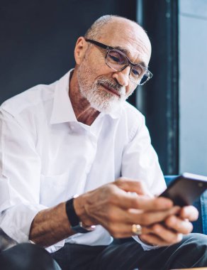 Gözlüklü sakin ve düşünceli bir girişimci rahat koltukta oturmuş sosyal ağda mesajlaşırken akıllı telefon ekranına bakıyor.