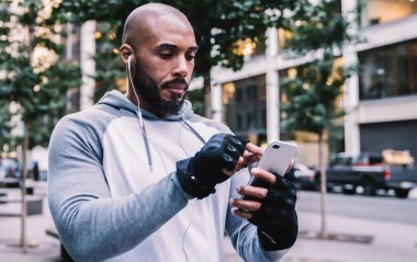 Sakallı, mesaj yazan ve müzik dinleyen siyah bir sporcu. Eğitimde ara verildiğinde kulaklık takıyor. Bulanık arka planda sokakta yürürken.