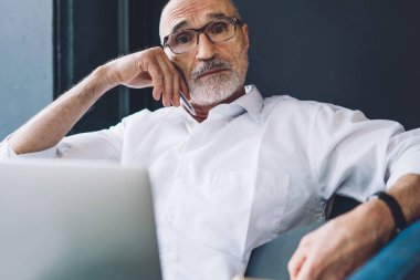 Beyaz tişörtlü, siyah pantolonlu, dizüstü bilgisayarla çalışan ve kameraya bakarken notlar alan yaşlı, sakallı, kel bir adamdan.