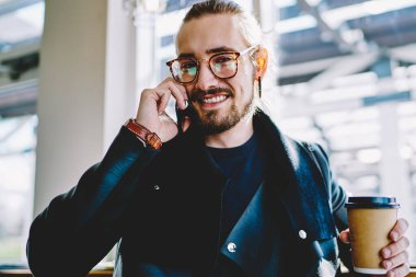 Kafeteryadaki kahve molası sırasında hücresel uygulamayla arkadaşça sohbet eden neşeli sakallı hippi adamın portresini kapat. Moda gözlüklü, kameraya gülümseyen ve telefonla arayan mutlu bir adam.