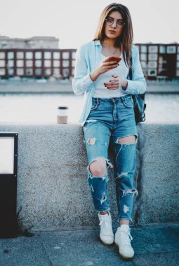 Kot pantolon giymiş genç bir kadının portresi, internet üzerinden mesajlaşırken kameraya bakarak kamuya açık 4G internet bağlantısını kullanıyor. Şehir merkezinde duran çekici hippi kız turist.