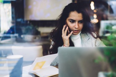 Uzun siyah saçlı, rahat giyimli, cep telefonuyla konuşan ve dizüstü bilgisayarla masada oturan zeki bir kadın.