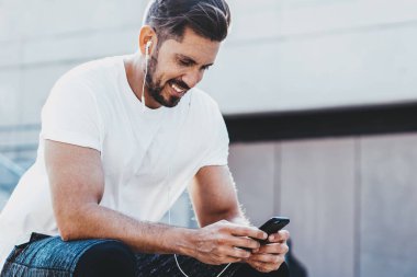 Aktif giyinmiş mutlu gülümseyen adam, dışarıda kardiyo eğitiminden sonra dinlenirken sağlıklı yaşam tarzı hakkında kitap okurken kulaklıklı neşeli bir adam, uygulamadan müzik dinlemekten keyif alıyor.