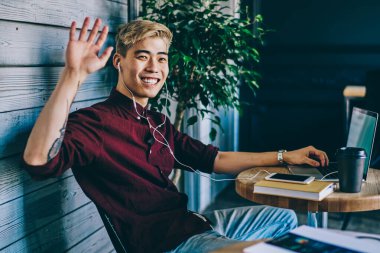 Uzaktan kumanda sırasında modern laptopa bağlı kulaklıkla müzik dinlerken el sallayan mutlu Çinli hipsterin portresi. İnternetteki yazılımı güncelleyen olumlu Japon genç adam.