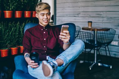 Modern kafenin içindeki koltukta oturmuş modern akıllı telefondan komik bir tebligat okurken elinde kahve tutan pozitif Çinli hippi blogcu. Neşeli Asyalı genç adam internette sohbet ediyor.