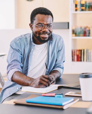 Gözlüklü ve günlük giysileri olan başarılı Afrikalı Amerikalı adam dizüstü bilgisayarla ışık tasarım ofisinde otururken not alıyor.