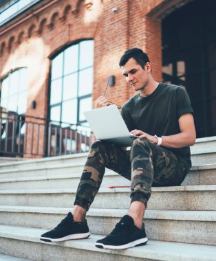 Ekrana odaklanan gözlüklü, askeri tarzda giyinmiş ve merdivenlerde otururken dizüstü bilgisayar kullanan düşünceli bir adamdan.