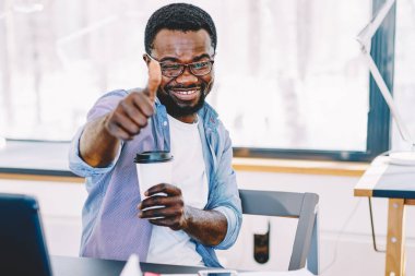Elinde bir bardak kahveyle ofiste otururken gülümseyen ve başparmağını gösteren Afro-Amerikalı bir adam.