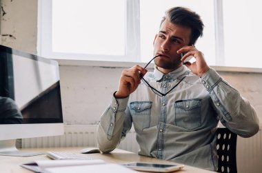 Sıradan giyinen düşünceli genç adam iş çözümünü düşünürken akıllı telefon aygıtında ofis bilgisayarında oturuyor. Telefonla düşünen bir yönetici arıyor.