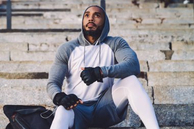 Genç Afrikalı Amerikalı atletik erkek taş merdivenlerde oturuyor ve kulaklıkla müzik dinliyor.