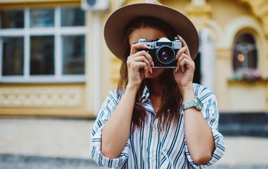 Sokakta fotoğraf çekmek için klasik kamera kullanan modaya uygun şapka ve giysili genç bir kadın, açık havada kentsel ortamların resmini yapan profesyonel bir kadın fotoğrafçı.