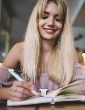 Not defterine yazarken gülümseyen uzun saçlı sarışın kadın ve rahat bir internet kafe projesinde dizüstü bilgisayar kullanıyor.