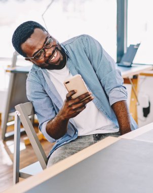 Gözlüklü neşeli siyah adam akıllı telefon ekranına bakıyor ve gülerek ofisteki masada oturuyor.