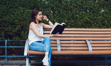 Kısa kahverengi saçlı, bacak bacak bacak üstüne atmış, sıradan giyinmiş, çitlere ve ağaçlara yaslanmış kitap okurken ve kafaya dokunurken konsantre olmuş Asyalı genç bayan.