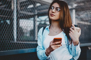 Gözlüklü genç bir kadının elinde lezzetli kahve içeceği ve akıllı telefon cihazıyla şehir ortamında dikilip düşünmesi. Düşünceli hippi blogcu yeni internet yayını için fikirler düşünüyor.