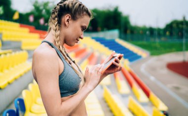 Spor giyimli sarışın atletik kadın ve cep telefonuyla mesajlaşan at kuyruklu, bulanık arka planda boş stadyum tribününde tek başına duran...
