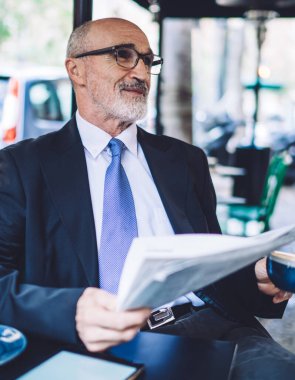 Caddedeki kafede gri sakallı, mavi kravatlı, kahve fincanı ve şehir arka planında gazetesi olan neşeli, son sınıf öğrencisi.