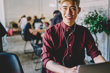 Modern laptop cihazına bağlı kulaklıkla müzik dinleyen olumlu Japon genç adam kablosuz internet kullanıyor. Yaratıcı Çinli hippi adam web sitesinden ses şarkıları dinlemekten hoşlanıyor.