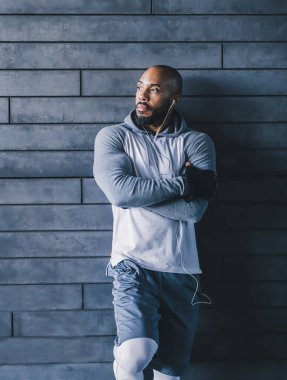 Beyaz kulaklıklı, beyaz kulaklıklı, duvara yaslanmış ve kollarını kavuşturarak fotokopi çekilen ve müziğin keyfini çıkaran kendine güvenen kel, kel sporcu.