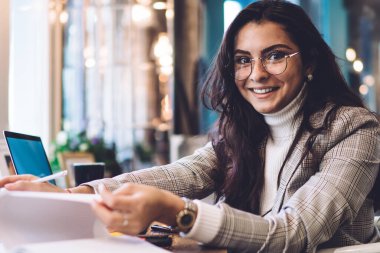 Gözlüklü, siyah saçlı, ceketli, zeki bir kadın kafeteryada dizüstü bilgisayarlı bir şekilde oturan defterin sayfalarını çeviriyor.