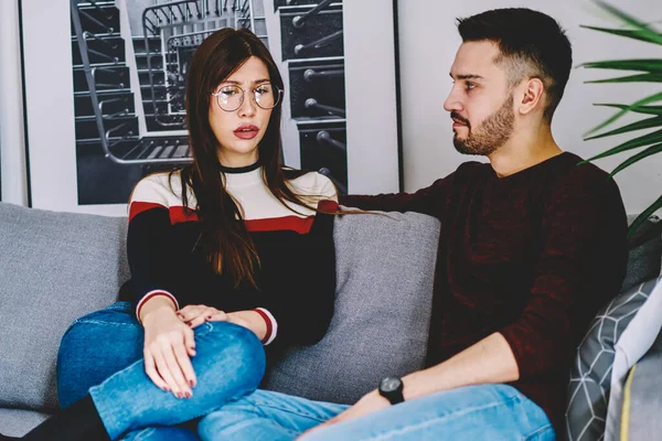 Serious wife and husband discussing problems about their marriage have quarrel with ways for solving while sitting on comfortable sofa in stylish home interior, couple in love communicating indoors