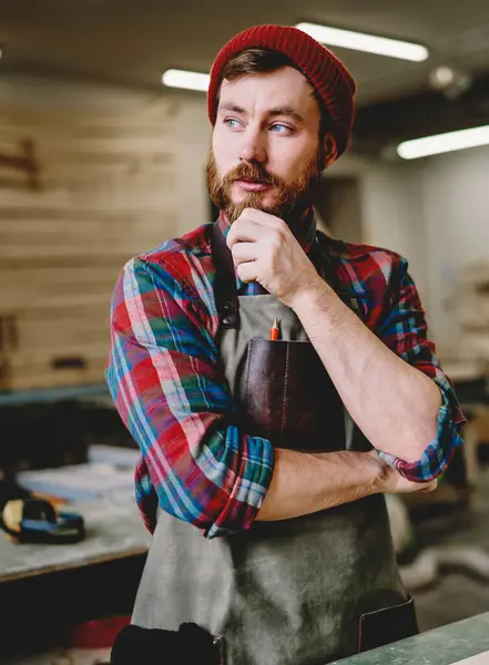 Bearded male carpenter in casual clothing hat and apron rubbing chin in though and looking away while working at modern workshop