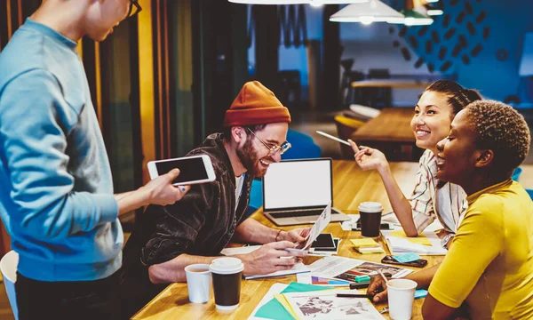 Happy crew of trendy dressed group of hipsters discussing ideas for startup sitting at meeting table with netbook and stationery, multiracial crew of students having brainstorming session together