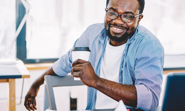 Content adult black man in glasses holding cup of coffee in hand and smiling at camera while sitting at laptop