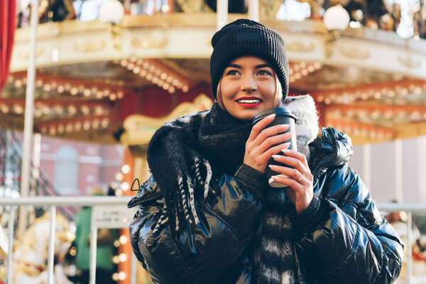 Toothy smiling young female with red lips wearing black winter hat standing by festive New Year carousel with coffee to go while dreaming and looking away