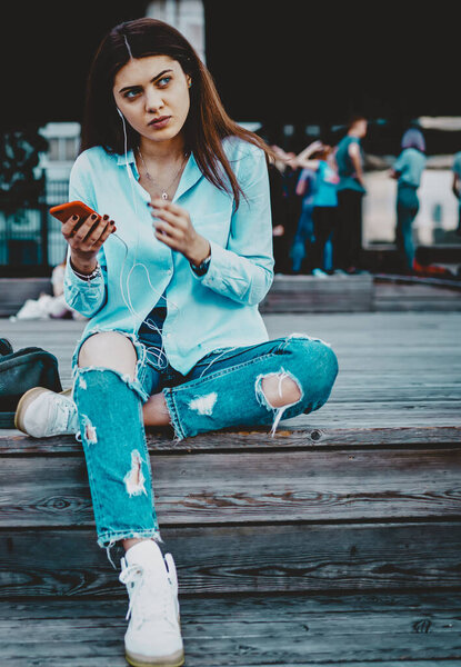 Обаятельная молодая женщина смотрит в сторону, сидя на улице и слушая музыку из скачанного плейлиста на смартфоне.