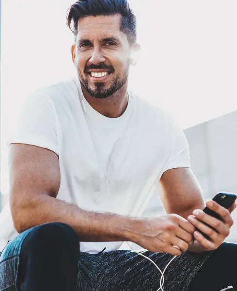 Portrait of cheerful male jogger sitting outdoors with cellular phone in hand and smiling at camera during time for training in city, happy man in headphones enjoying music sounds from website