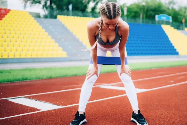 Young fit sportswoman standing bending forward with hands putted on knees while resting after doing squats with elastic band on outdoors stadium and looking down