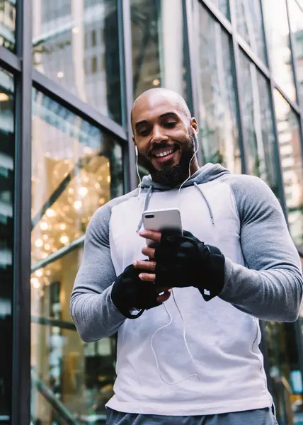 Low angle of smiling African American man in sportswear enjoying music and using smartphone while standing on street near glass office building