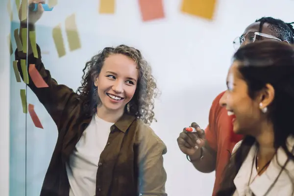 Charming curly woman with diverse group mates standing near glass wall and writing on colorful sticky notes while coworking on project