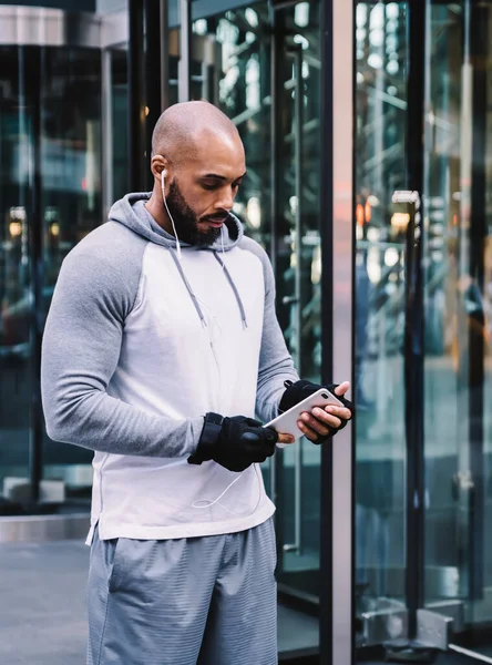 Bald African American man in sportswear using smartphone and listening to music while standing on street near door of modern office building