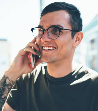 Gözlüklü ve dövmeli gülümseyen adam güneşli bir günde şehirde yürüyen arkadaşıyla sohbet etmek için telefonunu kullanıyor.