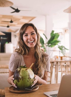 Bambu kafeteryasında egzotik tropikal içecekler içtiği için gülen neşeli Kafkasyalı serbest yazar portresi Endonezya hasır kafesinde kameraya gülümseyen mutlu kadın metin yazarı.