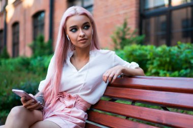 Beyaz tişörtlü, pembe şortlu, uzun pembe saçlı, cep telefonuyla müzik dinleyen caddedeki ahşap bankta oturan sıska kadın.