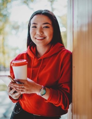 Gündelik giyinmiş neşeli Çinli kadının yarım boy portresi güneşli yaz günlerinde şehir ortamında vakit geçiriyor, kafein içeceği ve kameraya gülümseyen mobil aletli mutlu bir genç.