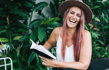 Çekici, şapkalı, boyalı saçlı, kitap okuyan, tropik çalıların yanında oturan ve kameraya bakan bir kadın.