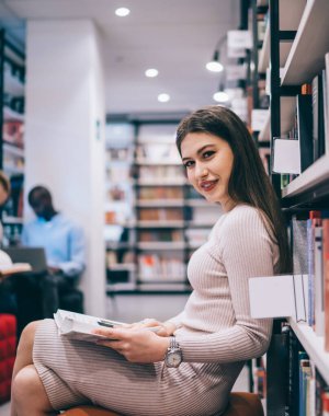 Çekici ve neşeli bir kadının kütüphanede bacak bacak bacak üstüne atmış kitap rafına yaslanmış, elinde kitap tutarken kameraya bakarken görüntüsü.